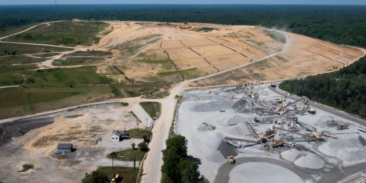 Aerial view of a landfill construction site with cleared land, earth-moving machinery, gravel piles, and a few small buildings, surrounded by trees and forested areas.
