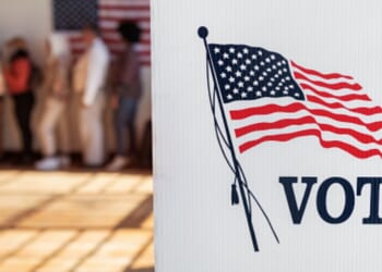 A voting booth stands in an auditorium with a line of citizens lined up behind it.