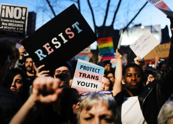 The World Professional Association for Transgender Health just made a key admission. WPATH said its Standards of Care are opinion, not objective fact claims. Pictured: a crowd of protesters holds signs, including RESIST, PROTECT TRANS YOUTH, and Spanish-language messages. People have raised fists, and a trees branches are visible in the background at dusk.
