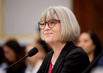 A woman with gray hair and glasses, wearing a black blazer and pearl necklace, speaks into a microphone at a formal event. Blurred audience members are seen in the background.