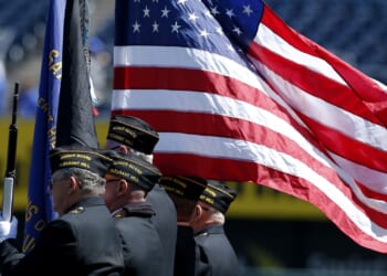 Three uniformed veterans stand in profile, carrying flags, including a large, unfurled American flag, during a ceremony at an outdoor event.