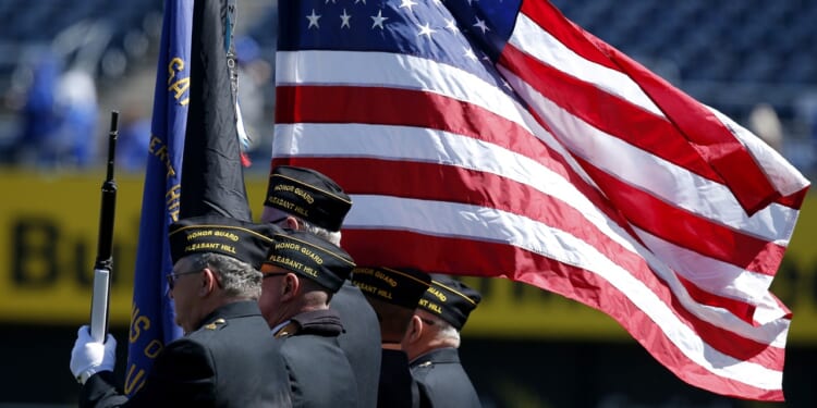 Three uniformed veterans stand in profile, carrying flags, including a large, unfurled American flag, during a ceremony at an outdoor event.