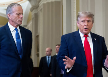 A man in a blue suit stands to the left while another man in a dark suit and red tie speaks with hands raised. They are indoors with blurred people and arched hallways in the background.