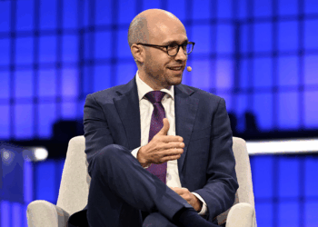A man in a suit and tie, wearing glasses and a headset microphone, sits onstage with a blue geometric background, gesturing with his hand while speaking.