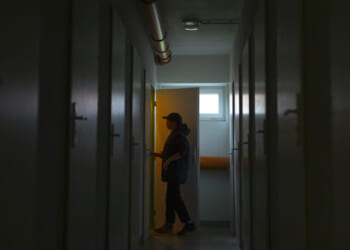 A person silhouetted in a dim hallway opens a door with warm light shining through, evoking a tense atmosphere. The hallway has closed doors on both sides and exposed pipes on the ceiling.