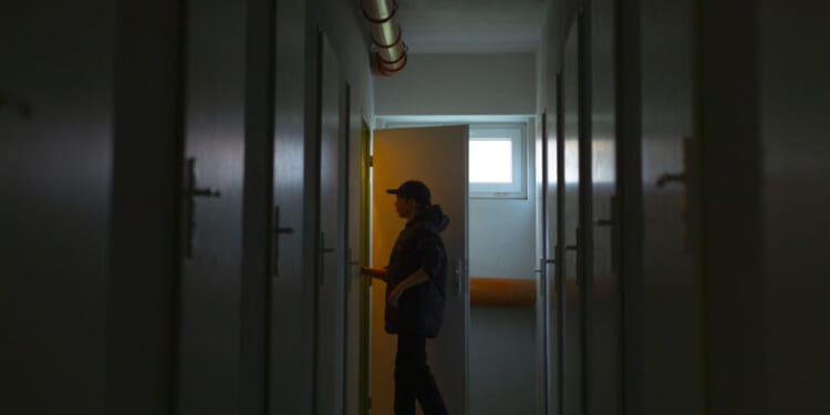 A person silhouetted in a dim hallway opens a door with warm light shining through, evoking a tense atmosphere. The hallway has closed doors on both sides and exposed pipes on the ceiling.