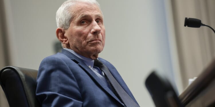 An older man in a blue suit and tie sits at a desk, looking slightly to his right. A microphone is positioned on the desk in front of him. The background is out of focus.