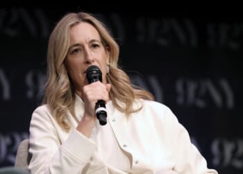 A woman with long blonde hair speaks into a microphone while seated on stage. She wears a white jacket and a neutral expression. A dark backdrop with 92Y in light letters is behind her.