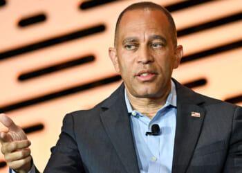 Hakeem Jeffries, wearing a suit, gestures with his left hand while speaking, seated in front of a blurred background with horizontal black and orange patterns.