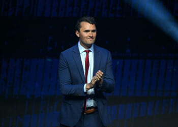 Charlie Kirk, dressed in a blue suit and red tie, stands on stage, clapping his hands, with a dark blue background and a spotlight shining from the upper right corner.