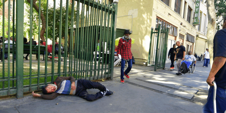 A person lies on the sidewalk by a green fence, while several people walk nearby. A building with large windows and a hotel sign is in the background. Trees and benches are visible inside the fenced area.