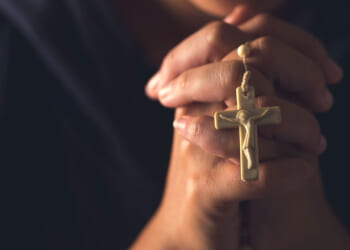 Hands clasped in prayer holding a gold crucifix necklace, with a blurred background.