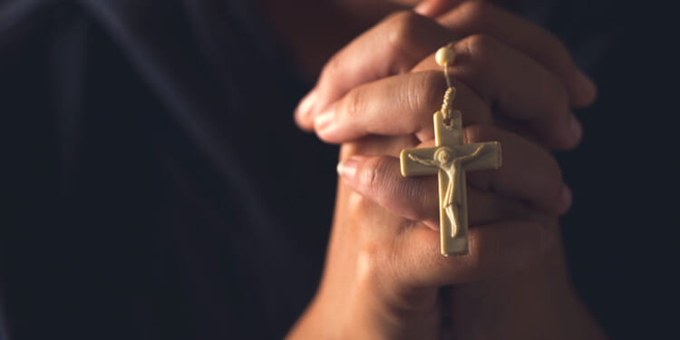 Hands clasped in prayer holding a gold crucifix necklace, with a blurred background.