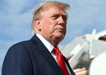 President Donald Trump in a suit and red tie stands outdoors with an airplane staircase in the background and a blue sky overhead.