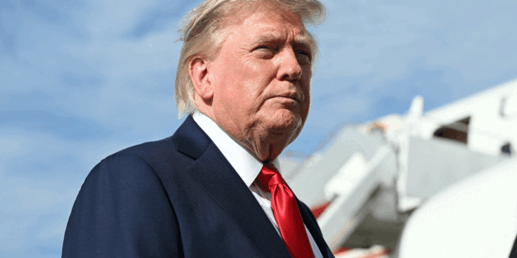 President Donald Trump in a suit and red tie stands outdoors with an airplane staircase in the background and a blue sky overhead.