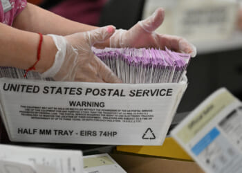 A person wearing gloves handles a USPS tray filled with mail-in ballots. The tray is labeled HALF MM TRAY – EIRS 74HP. Other paperwork is visible in the foreground.