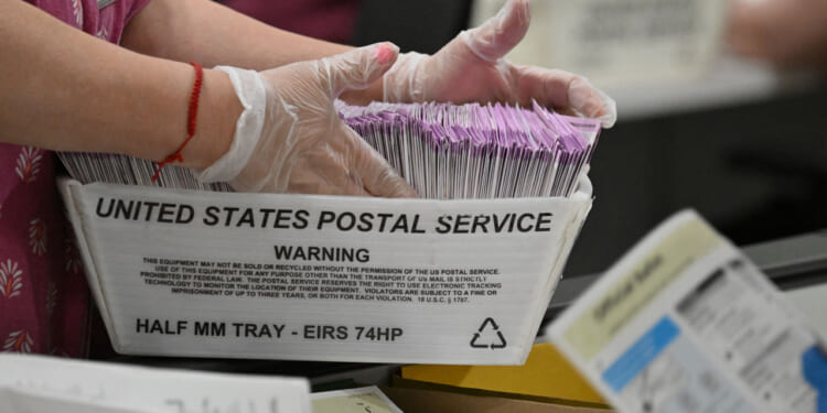 A person wearing gloves handles a USPS tray filled with mail-in ballots. The tray is labeled HALF MM TRAY – EIRS 74HP. Other paperwork is visible in the foreground.