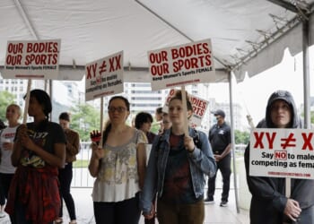 People hold signs reading OUR BODIES, OUR SPORTS and XY ≠ XX at an outdoor protest under a canopy. The group appears focused and engaged, standing together in an urban setting.