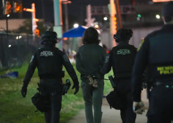 Three police officers escort a handcuffed person at night near a fence; the officers wear tactical gear labeled Newark Police..