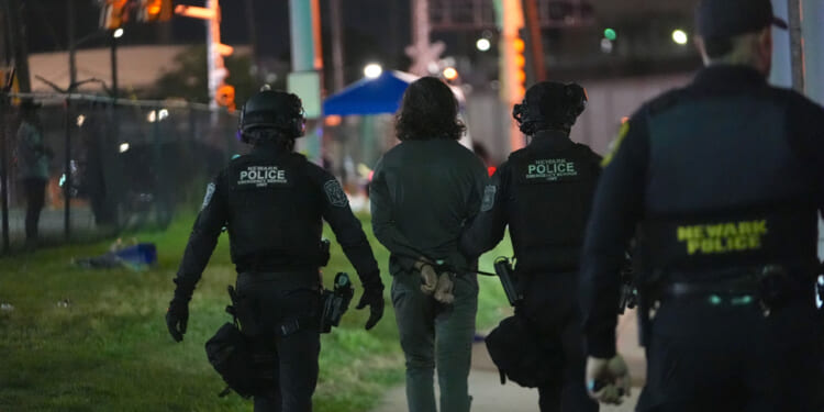 Three police officers escort a handcuffed person at night near a fence; the officers wear tactical gear labeled Newark Police..