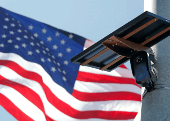 A security camera mounted on a pole with a solar panel attached, with a large American flag waving in the background against a clear blue sky.