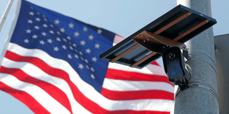 A security camera mounted on a pole with a solar panel attached, with a large American flag waving in the background against a clear blue sky.