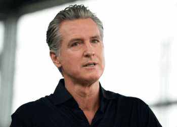 A man with gray hair, wearing a dark collared shirt, speaks while looking slightly off camera. The background is blurred, suggesting an indoor or semi-outdoor setting.