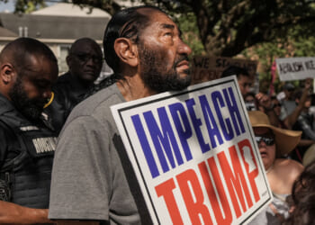 A man holds an Impeach Trump sign at a protest, surrounded by people and police officers. Some hold signs, and trees and houses are visible in the background.