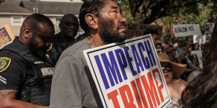 A man holds an Impeach Trump sign at a protest, surrounded by people and police officers. Some hold signs, and trees and houses are visible in the background.