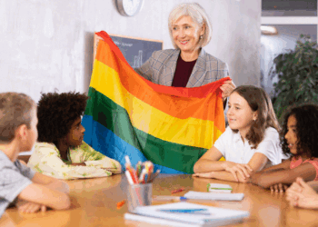 An older woman holds a rainbow pride flag and smiles while sitting at a table with five children. The children are listening and looking at the flag in a classroom setting.
