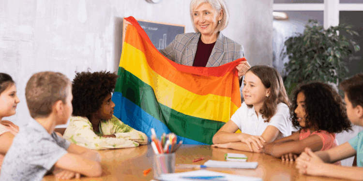 An older woman holds a rainbow pride flag and smiles while sitting at a table with five children. The children are listening and looking at the flag in a classroom setting.