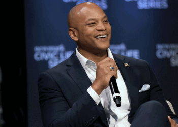 Maryland Gov. Wes Moore in a dark suit holds a microphone while seated on stage, speaking at an event with a Civic Life Series banner in the background.
