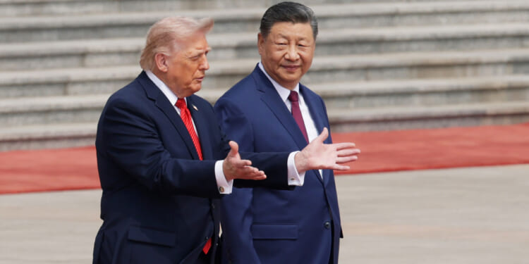Two men in suits, one gesturing with open hands while speaking and the other looking ahead and smiling, stand outdoors on a wide stone plaza in China, with steps and a red carpet in the background.