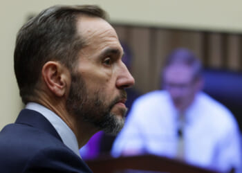 A man in a suit with a short beard listens intently during a meeting, while a blurry figure appears on a screen in the background.