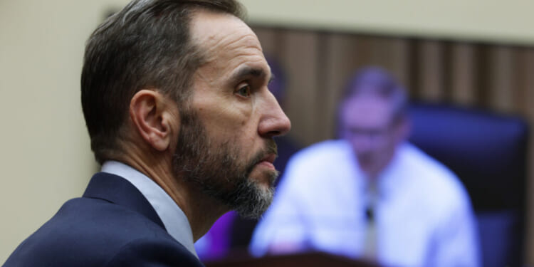 A man in a suit with a short beard listens intently during a meeting, while a blurry figure appears on a screen in the background.