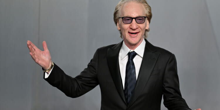 A man in a black suit, white shirt, and dark tie smiles and gestures with one hand raised. He has light hair, wears glasses, and stands against a plain gray background.