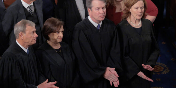 Four judges in black robes stand side by side in a courtroom, with others seated and standing around them. The judges have serious expressions and their hands are clasped or held together in front of them.