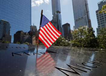 An American flag stands on the engraved names at the 9/11 Memorial in New York City, with tall buildings and trees in the background on a sunny day.