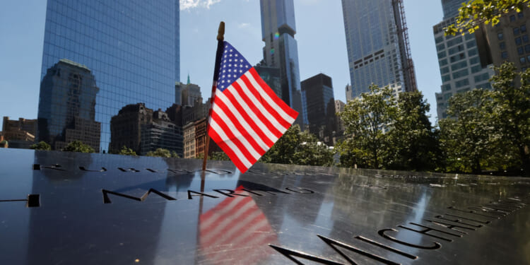 An American flag stands on the engraved names at the 9/11 Memorial in New York City, with tall buildings and trees in the background on a sunny day.