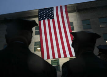 Silhouetted figures in uniform stand facing a large American flag hanging on the side of a building at dusk.