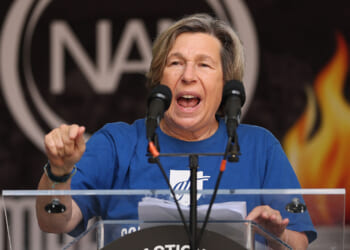 AFT's Randi Weingarten is influencing AI through corporate partnerships. Pictured: a person wearing a blue shirt speaks passionately at a podium with two microphones, gesturing with one hand. A blurred logo and flames are visible in the background.