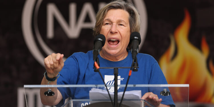AFT's Randi Weingarten is influencing AI through corporate partnerships. Pictured: a person wearing a blue shirt speaks passionately at a podium with two microphones, gesturing with one hand. A blurred logo and flames are visible in the background.