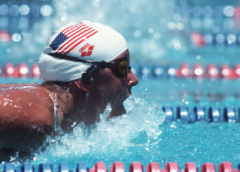 A swimmer wearing a white USA swim cap and goggles competes in a race, emerging from the blue water with an open mouth, creating splashes, surrounded by red, white, and blue lane dividers.