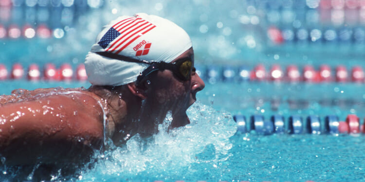 A swimmer wearing a white USA swim cap and goggles competes in a race, emerging from the blue water with an open mouth, creating splashes, surrounded by red, white, and blue lane dividers.