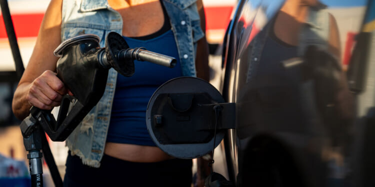 A person in a denim vest and blue top holds a diesel gas pump nozzle, preparing to refuel a dark-colored vehicle at a gas station.
