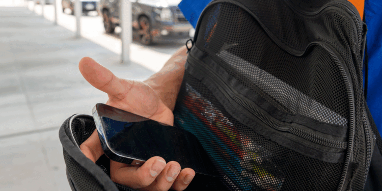 A student holds a smartphone device while opening the mesh pocket of a black backpack outdoors.