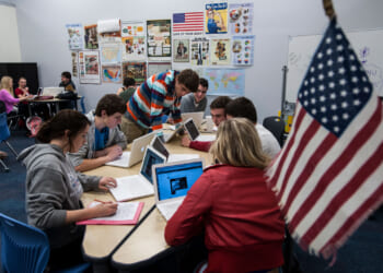Students sit at tables in a classroom, using laptops for schoolwork. Educational posters and maps—including references to Western civilization—decorate the walls. An American flag is in the foreground. Small groups work together, engaging with their computers and each other.
