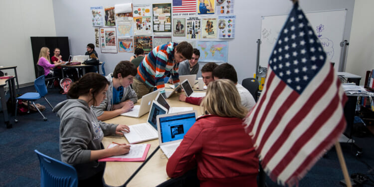 Students sit at tables in a classroom, using laptops for schoolwork. Educational posters and maps—including references to Western civilization—decorate the walls. An American flag is in the foreground. Small groups work together, engaging with their computers and each other.