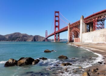 The Golden Gate Bridge stretches over blue water with waves splashing on a sandy, rocky shore under a clear sky. Hills are visible in the background.