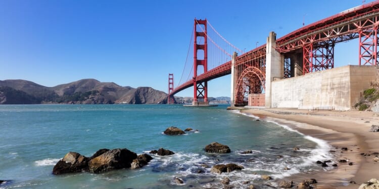 The Golden Gate Bridge stretches over blue water with waves splashing on a sandy, rocky shore under a clear sky. Hills are visible in the background.
